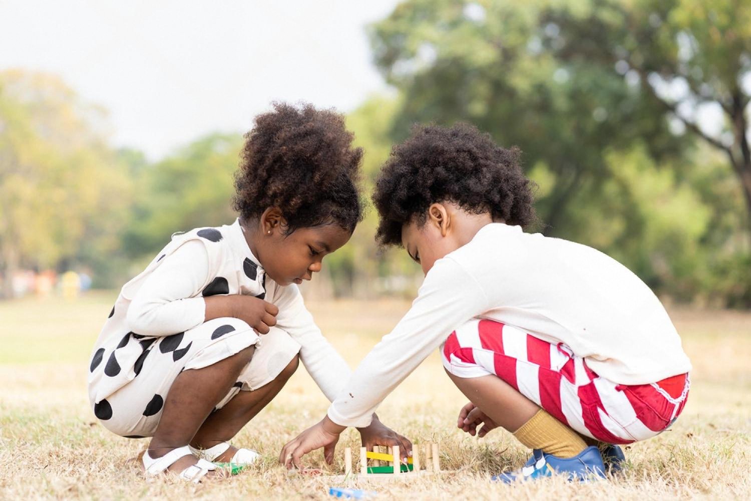 Two young children crouching on grass playing with a small wooden travel toy set